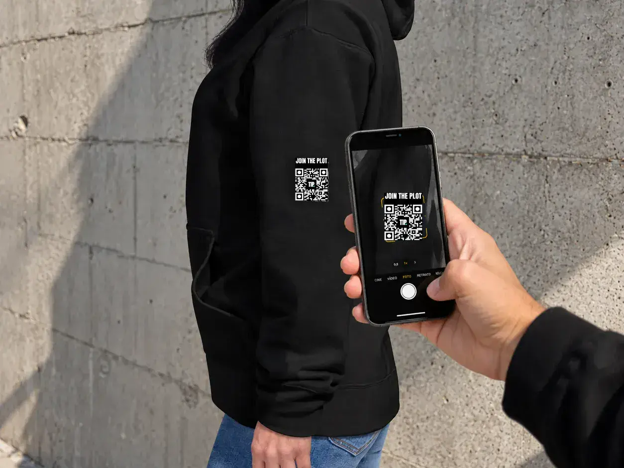 The Invisible Plot_DE 4 Eine Person scannt mit dem Smartphone den QR-Code „Join the Plot“ auf dem Ärmel eines The-Invisible-Plot-Hoodies
